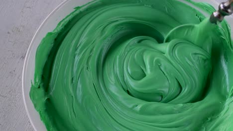 mixing-light-green-buttercream-icing-in-bowl-with-spoon-on-white-background-top-view