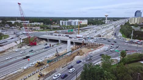 Slow-panning-rising-aerial-drone-shot-of-busy-highway-exit-intersection-with-overpass-and-road-construction-zone-showing-vehicular-traffic,-road-construction-equipment,-and-buildings-in-background