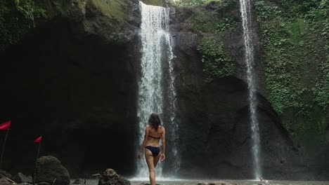 A-young-girl-slowly-walking-to-the-waterfall-and-looking-at-it