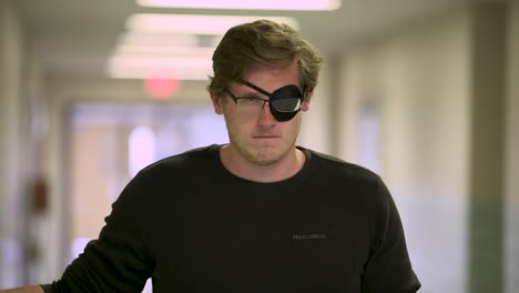 White-Man-with-Eye-Patch-and-Glasses-Stares-at-Camera-Seriously-in-a-Hallway