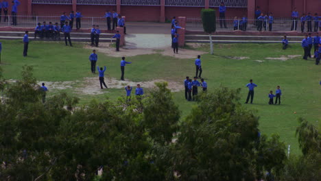 School-students-playing-in-the-ground-aerial-view-passing-the-trees,-students-are-in-blue-pants-and-shirts,-some-part-is-missing-of-grass-in-the-ground,-school-building-in-the-background