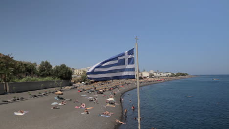 Weitwinkelaufnahme-Des-Kamari-Strandes-Auf-Santorini-Mit-Einer-Flatternden-Griechischen-Flagge-Im-Vordergrund