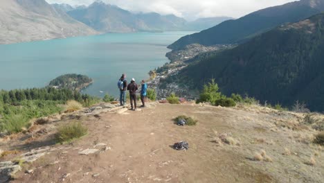 SLOWMO---Three-young-men-travellers-standing-on-Queenstown-Hill-track-hike,-New-Zealand-and-looking-at-the-Lake-Wakatipu,-mountains-with-fresh-snow-and-clouds---Aerial-Drone