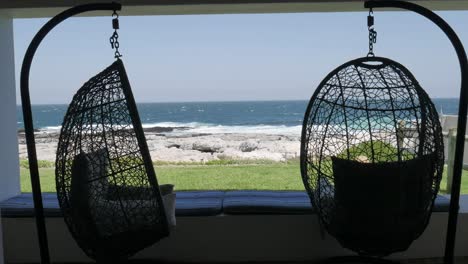 Hanging-chairs-on-verandah-swinging-gently-in-ocean-breeze-with-lovely-ocean-views-in-background-in-Hermanus,-South-Africa