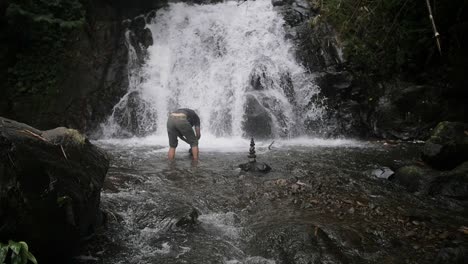 Zeitlupenaufnahme-Eines-Indonesischen-Mannes,-Der-In-Einem-Fluss-Vor-Einem-Wasserfall-Steinhaufen-Baut