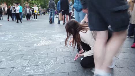 Two-girls-and-other-tourists-drawing-chalk-art-on-busy-pavement-in-London,-Leicester-Square,-England,-UK,-Europe