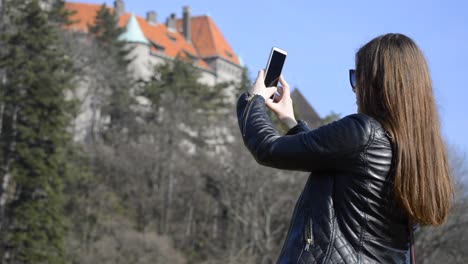 A-lovely-young-woman-in-black-leather-jacket-taking-pictures-of-a-castle-on-her-phone-in-a-public-park-on-a-nice-spring-sunny-day