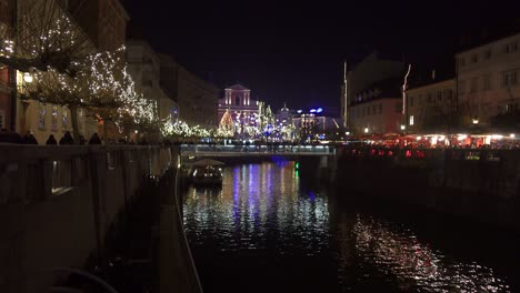 People-walk-along-a-river-illuminated-by-Christmas-lights-in-Ljubljana,-Slovenia