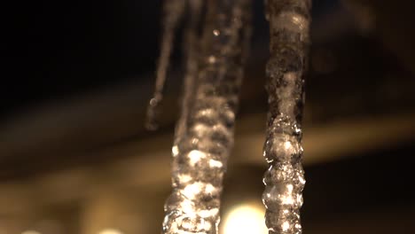 Backlit-Melting-Icicle-hanging-from-roof,-dripping-in-slow-motion,-shot-panning-downwards