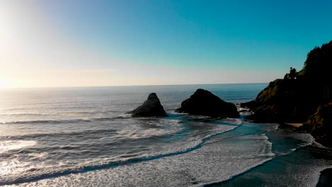 Beautiful-blue-calm-waves-slowly-rolling-onto-the-beach-along-the-coast-of-Oregon-and-the-Pacific-Ocean-with-blue-skies-and-silhouetted-rocks-in-the-background