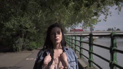 Portrait-of-an-attractive-latina-tourist-with-black-wavy-hair-and-a-jean-jacket-walking-in-a-park-in-London-with-a-view-of-Putney-bridge-behind-her