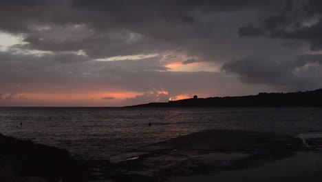 sunrise-timelapse-in-Qawra,-Malta,-sun-raising-up-behind-very-dark-rainy-stormy-clouds,-and-rain-start-to-fall