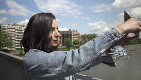 Attractive-Hispanic-tourist-taking-a-picture-of-London-from-a-bridge,-taking-her-phone-out-and-holding-it-to-frame-the-shot