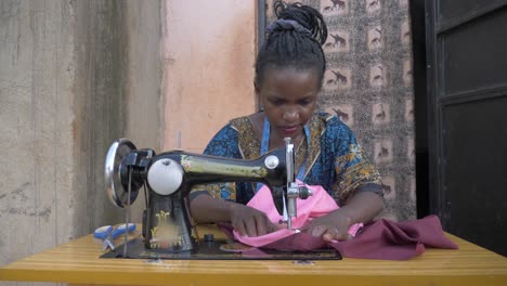 Front-shot-of-an-African-woman-sewing-cloths-on-a-tailoring-machine-in-Africa-outside-her-small-home-in-slow-motion