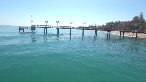 drone-flying-under-pier,-dangerous-risky-flight-ona-beautiful-sunny-beach-in-marbella,-malaga,-spain,-costa-del-sol