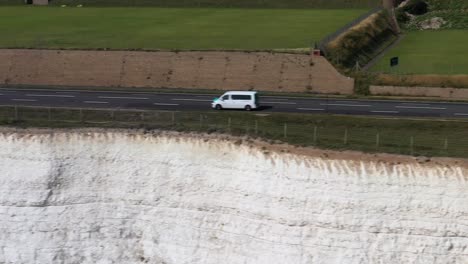 Aerial-tracking-shot-of-a-white-camper-van-on-the-A259-coast-road,-above-chalk-clifftops-near-Brighton,-UK