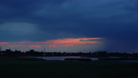 Time-lapse-of-dramatic-dark-thunderstorm-clouds-with-lightning-strikes-rolling-over-the-city-and-lake-Liepaja-after-the-sunset,-wide-shot