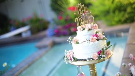 Shot-of-a-beautifully-decorated-floral-wedding-cake-sitting-on-a-pedestal-right-next-to-a-pool-during-a-summer-wedding-reception