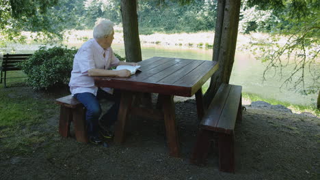 Older-man-reads-book-at-wooden-table-under-trees-near-river