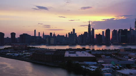 Dramatisches-Abheben-Der-Drohnenkamera-Aus-Der-Luft-–-über-Wasser-Schwebend,-Mit-Der-Ostseite-Von-New-York-City-Im-Hintergrund-–-Ein-Violetter-Sonnenuntergangshimmel