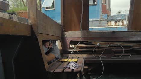 Young-man-wearing-mask-painting-underside-of-deck-interior-wooden-boat-cabin