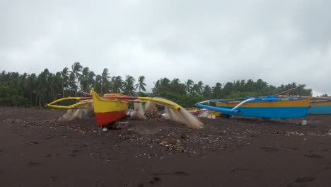Nahaufnahme-Von-Fischerbooten-An-Einem-Dunklen-Tropischen-Sandstrand-Mit-Bewölktem-Himmel,-Während-Ein-Windiger-Sturm-Palmen-Auf-Den-Philippinen-Weht,-4k