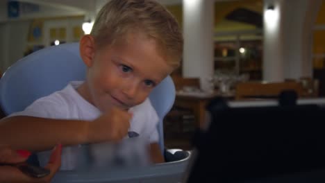 Three-year-old-child-sitting-at-table-at-restaurant,-eats,-laughs-and-looks-at-the-tablet,-family-concept