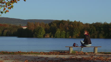 Young-woman-walks-up-and-sits-on-bench-at-lake-with-nice-view,-static