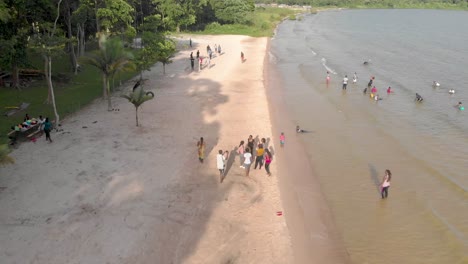 Aerial-shot-moving-forward-and-angled-down-at-African-youth-on-a-crowded-sandy-beach-on-the-shores-of-Lake-Victoria-in-Uganda-with-people-swimming-and-playing-sport