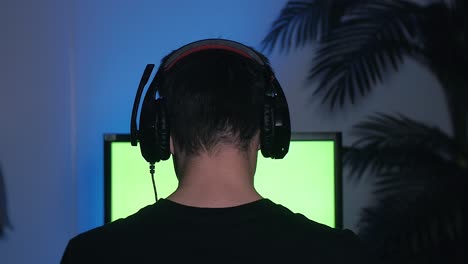 Gamer-Puts-His-Headset-with-a-Mic-On-and-Starts-Playing-computer-Games-on-green-screen-monitor