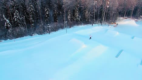 Side-aerial-view-of-a-snowboarder-jumping-off-a-slope-in-a-terrain-park-in-the-evening