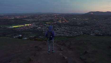 Dolly-follow-shot-of-girl-walking-down-the-Arthurs-seat-mountain-on-the-rocky-hiking-trail-in-evening,-dusk-with-city-of-Edinburgh-in-the-background-during-wonderul-blue-hour