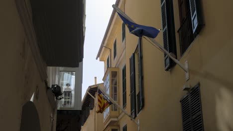 Europäische-Flagge-Und-Katalonien-Flagge-Winken-Auf-Einem-Fahnenmast,-Der-An-Einem-Gebäude-Unter-Einem-Fenster-In-Einer-Engen-Gasse-Befestigt-Ist