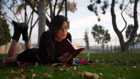 A-young-adult-woman-reading-a-novel-or-story-book-in-the-park-at-sunset-laying-in-the-grass-with-autumn-leaves-SLOW-MOTION