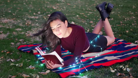An-attractive-young-woman-reading-a-story-book-or-novel-in-a-park-at-twilight-with-lights-glowing-around-her