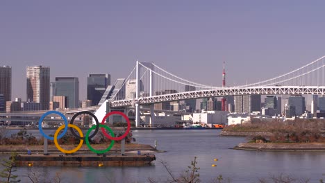 Señal-De-Anillo-De-Los-Juegos-Olímpicos-En-La-Bahía-De-Tokio-Con-La-Torre-De-Tokio-Y-El-Puente-Del-Arco-Iris-En-Un-Día-Brillante-Y-Soleado