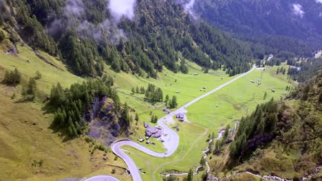 Winding-road-along-Dolomite-mountain-pass-in-the-Italian-Alps-with-a-water-stream-and-lodge-along-the-way,-Drone-flyover-reveal-shot