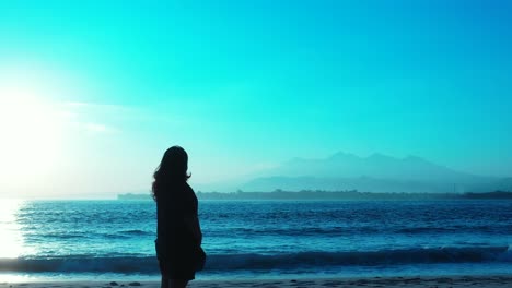 Lonely-girl-standing-in-front-of-blue-seascape-with-sea-waves-splashing-on-exotic-beach-of-tropical-island-on-a-bright-blue-sky-background,-Vietnam