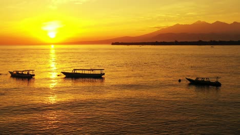 Silhouette-Von-Booten,-Die-Auf-Einer-Ruhigen-Meeresoberfläche-Bei-Goldenem-Sonnenuntergang-Schwimmen,-Mit-Gelber-Sonne,-Die-Am-Horizont-Der-Berge-In-Indonesien-Untergeht