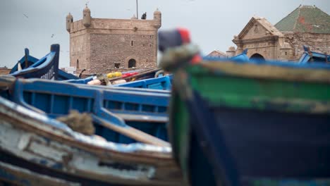 Die-Kamera-Bewegt-Sich-Nach-Links,-Mit-Verschwommenen-Fischerbooten-Im-Vordergrund-Und-Einer-Festung-Und-Hafeneinfahrt-Im-Fokus-In-Essaouira,-Marokko