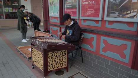 A-little-boy-and-a-mother-carrying-a-baby-give-coins-to-an-old-asian-street-musician-playing-a-Yangqin-or-chinese-hammered-dulcimer-at-Cabramatta