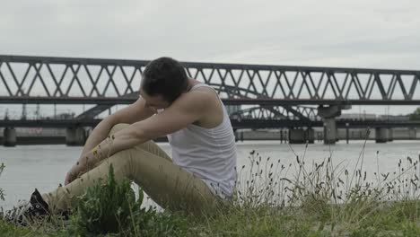 Side-view-of-positive-young-male-in-casual-outfit-rotating-head-and-looking-at-camera-with-smile-while-sitting-against-modern-urban-bridge-crossing-river