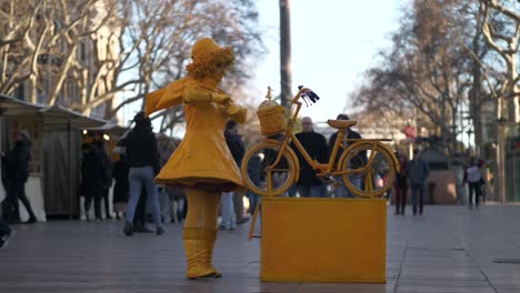 Slomo-of-female-street-performer-in-yellow-dress-and-hat-with-bike-in-Barcelona,-pedestrians-walk-by