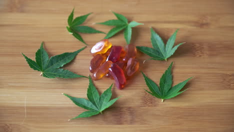 Push-in-to-CBD-Gummies-in-Pile-Surrounded-by-Circle-of-Marijuana-Leaves-on-Light-Wood-Tabletop
