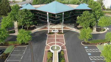 Modern-glass-office-building-in-business-park,-aerial-push-in-shot-during-summer,-blue-green-teal-metal-roof,-geometric-architecture-design,-beautiful-landscaping,-establishing-shot