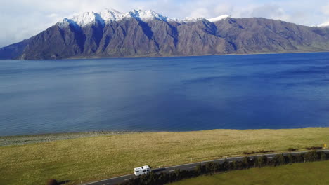 Aerial-video-of-a-Van-driving-on-a-Coastal-road-with-green-paddocks,-then-panning-to-the-blue-ocean,-and-Snowy-mountains