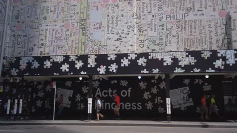 People-walking-on-a-sidewalk-in-Sydney-CBD-downtown-shopping-district-with-the-Act-of-Kindness-writing-with-map-of-Sydney-city-building-art-above-them