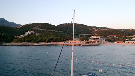 Eine-Luxuriöse-Weiße-Yacht-Mit-Einem-Hohen-Mast-Ohne-Segel,-Die-Am-Abend-Eines-Sonnigen-Sommertages-In-Der-Nähe-Des-Strandes-Der-Küstenstadt-Utjeha,-Montenegro,-Langsam-Auf-Den-Wellen-Schaukelt,-Luftaufnahme