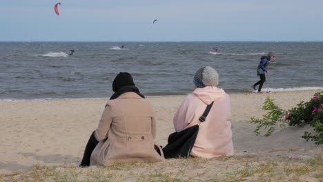 Two-Persons-Sitting-In-The-Sand-Watching-The-Kitesurfers-On-The-Baltic-Sea-In-Rewa,-Poland---midshot