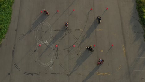 top-down-wide-aerial-view-of-multiple-riders-practicing-on-an-advanced-motorcycle-training-slalom-course-between-orange-cones-with-long-shadows,-and-tire-marks